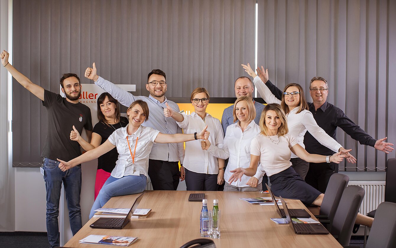 Teamfoto von Mitarbeitenden des Faller Packaging Standorts Łódź – motiviertes Team in fröhlicher Pose im Besprechungsraum.